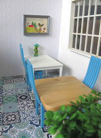Modern dolls house miniature cafe scene, with blue and white tiled floor, white on white wallpaper, white and beech tables, blue chairs and plants in the foreground.