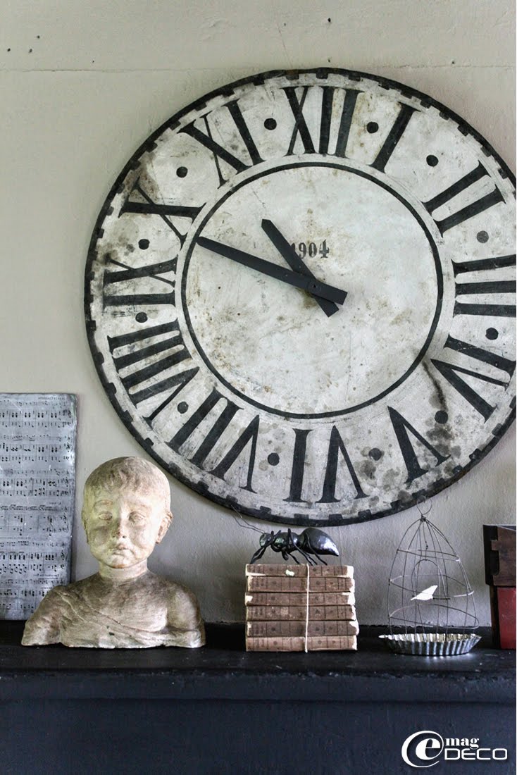 Posés sur la cheminée de la cuisine, un ancien cadran d’horloge, un buste, une pile de livres chinés et une fourmi en papier mâché, création Farfelus Farfadets
