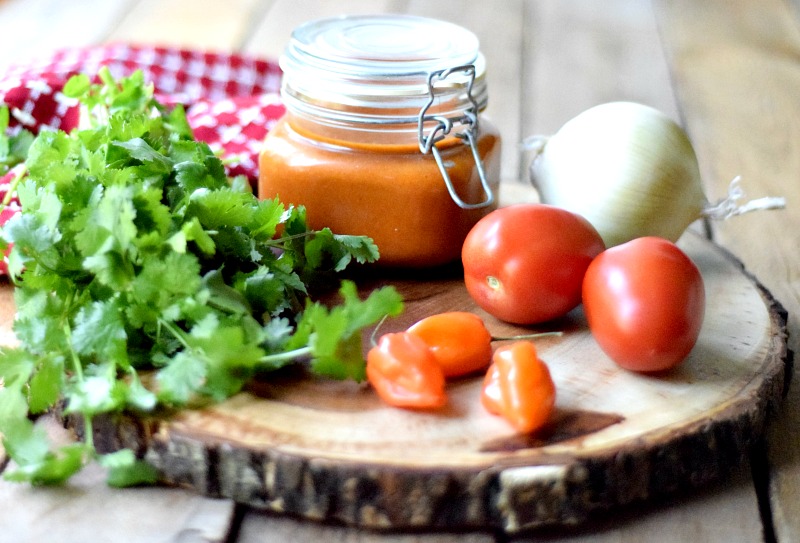 Chile de Arbol Hot Sauce in a glass jar on a round wooden cutting board with onion, tomato, habanero, and cilantro