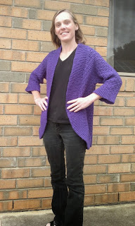 Jodie modelling the purple Belcarra cardigan over black tee shirt and black jeans. The cardigan has three-quarter length slightly flared sleeves with ribbing around the curved hem, front edges and shawl collar. The front corners of the bottom hem are curved.