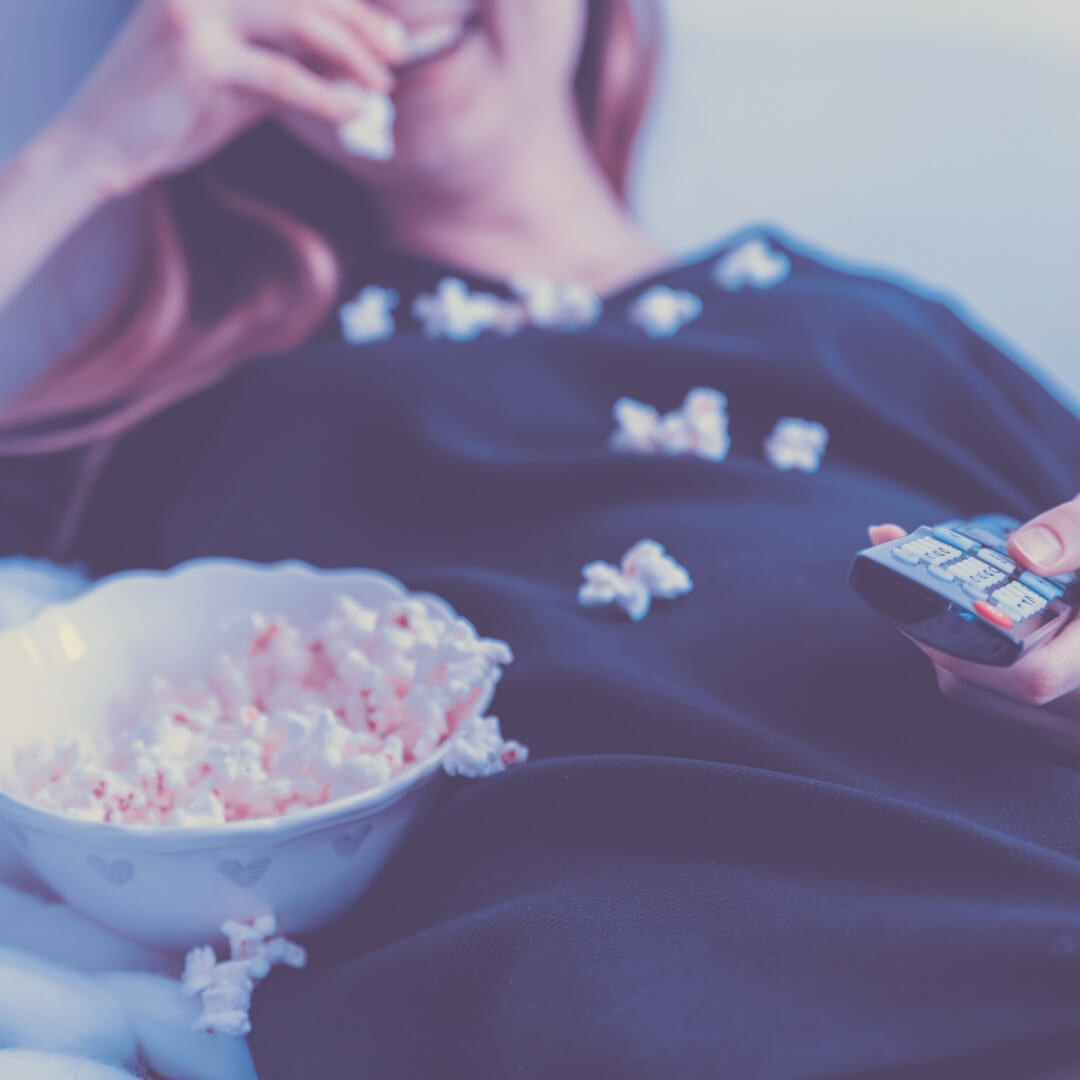 A woman sits with a bowl of popcorn resting next to her. She is holding a remote for the TV and popcorn kernels rest on her body. The camera is focussed on the popcorn. Image for The best movies to watch with your children.