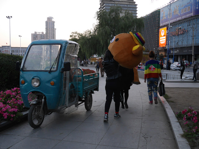 young woman carrying a bear mascot head past a three-wheeled vehicle in Mudanjiang, China
