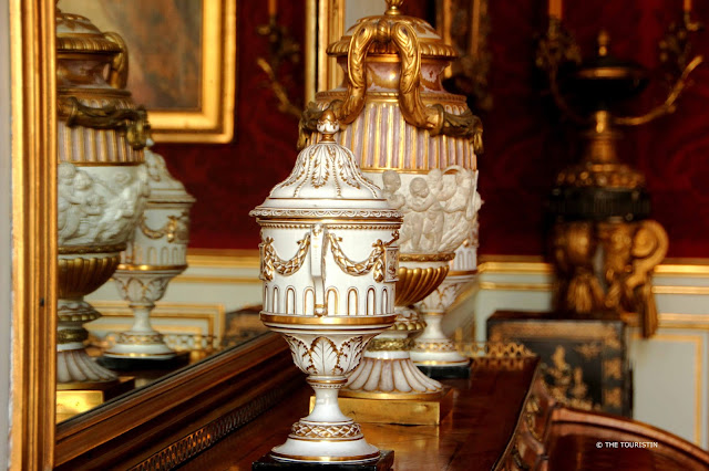 Two white gold-ornamented porcelain urns on a mantel frame in front of a mirror.