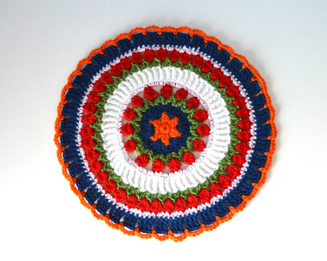 Mandala has an orange star in the centre surrounded by a blue circle. Two concentric rings of green 'leaves' and red 'tulip flowers' are separated by a wider ring of tall white stitches. After the second ring of tulips is a thin stripe of white double crochets, ending with a wider blue border with orange chain loops to finish. 