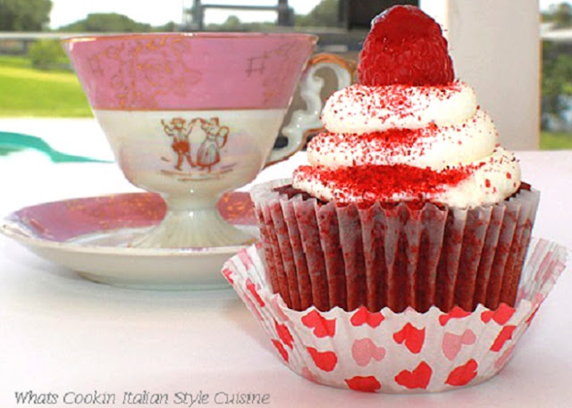 Raspberry Red Velvet Cupcakes this photo is of two cupcakes that are raspberry and red velvet. They have whole raspberries on top and cream cheese frosting in heart paper liners