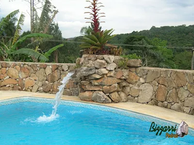 Construção de piscina com muro de pedra, a cascata de pedra com a bica d'água de madeira e o piso com caco de pedra São Tomé.