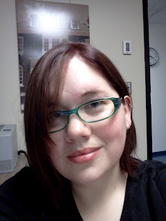 Self-portrait on my phone. Picture shows me, wtih dark red hair, green glasses, black shirt. Background is a white wall with framed picture of a campus building.