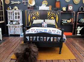 One-twelfth scale miniature scene, with a bed dressed in linen and black with throw rug and cushion printed with letter As, in front of a false wall. To the left of the bed is a distressed dresser and to the right is a set of pipe shelves.