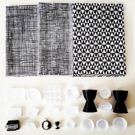 Flat lay of black and white items including fabric pieces, a one-twelfth scale modern miniature toaster, white platters, black Philippe Starck Prince Aha stools, a cameo and a black wine goblet