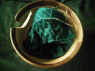 A lace leaf stitch pattern on circular needles. Yarn is a deep turquoise/teal with a cabled texture.  Knitting and yarn are both in a wooden yarn bowl