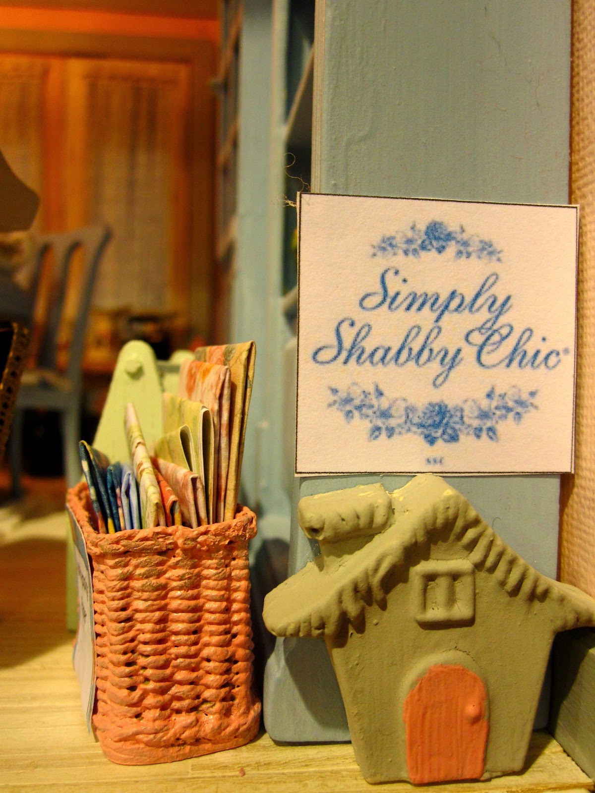 Modern miniature shabby chic shop bookcase with 'Simply shabby chic' sigh, pink painted basket filled with paper and a small model house. 