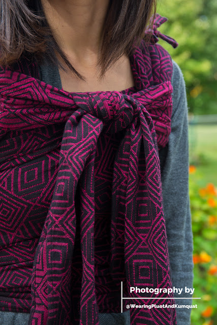 [Image of a tan skin Asian woman with dark brown hair wearing a not visible sleeping toddler on her back in a magenta and black concentric square patterned meh dai baby carrier. Woman's face isn't visible. Image is from neck through torso. The carrier covers her front torso with a double knot at the top of her chest. Bottom right corner in white is a watermark with a "T" lying on its side; top text "Photography by," bottom text "@WearingPluotAndKumquat."]