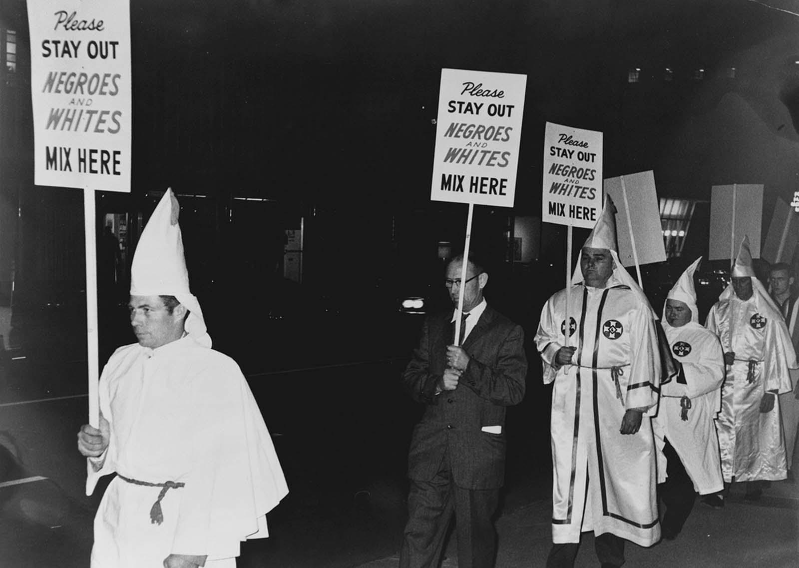 Several Ku Klux Klan members, most in white robes and unmasked hoods, carry placards stating: 