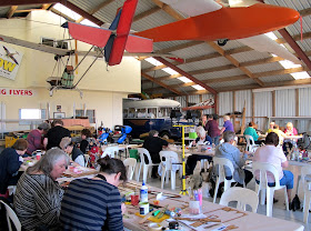 Group of people doing workshops in a hangar, with a bus in the far corner and a plane hanging above them.