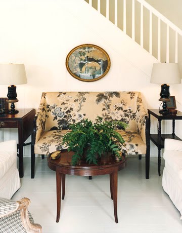 Living room in a colonial home with a chintz English settee between two black side tables with a round, wooden coffee table in front