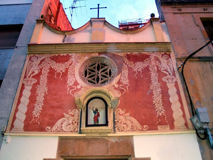Detall de la façana principal de la capella de Sant Joan decorada modernament amb esgrafiats de l'artista local Jaume Amat. Podem observar la fornícula amb la imatge de Sant Joan Baptista, i una rosassa amb una flor de sis pètals