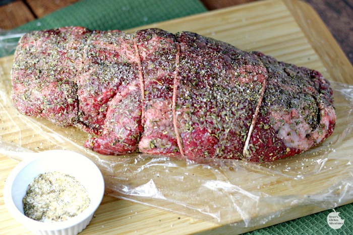 Garlic Herb Beef Tenderloin Roast with Creamy Horseradish Sauce by Renee's Kitchen Adventures raw on wooden cutting board with small white bowl of seasonings and roast seasoned and wrapped in plastic wrap  