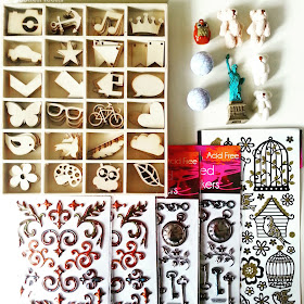Selection of wooden crafting shapes, dimensional stickers, miniature teddy bears, marble-effect beads plus an owl-shaped bell and miniature Statue of Liberty set out on a desktop.