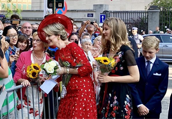 Crown Princess Elisabeth is wearing black floral print wrap midi dress. Queen Mathilde wore a red lace dress by Natan. Princess Eleonore