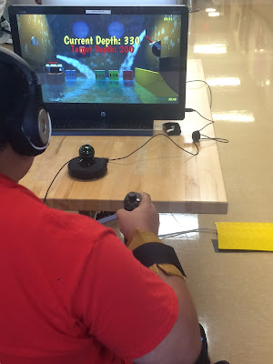 A photo from behind a patient as they wear headphones and hold a controller in their right hand while looking at a screen. In front of the screen is a motion tracker.