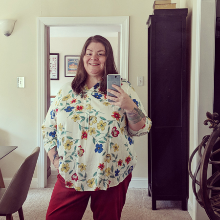 image of me standing in a mirror, wearing a white blouse with a colorful floral pattern and red jeans