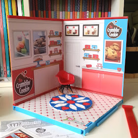 MiWorld cookie shop walls and floor assembled, with a miniature red Eames rocking chair and a round retro rug in colours of red, teal and white on the floor.