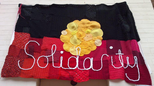 A large patchwork Australian Aboriginal flag in black, yellow and red with the word 'Solidarity' in white across the red strip.