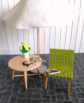 Modern dolls' house miniature patio with market umbrella, mid-century woven chair with a magazine and table with a glass of water, sunglasses and a fan on it.