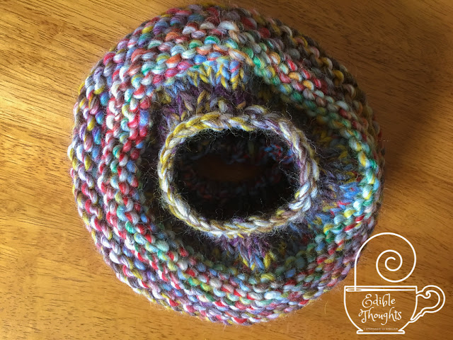 [Image of the top of a multi-colored chunky wool yarn knit hat. Focused on the hole on top for a messy hair bun. The hat lays on a wood surface. Bottom right corner watermark in white is a tea cup with a needle lying horizontally across the rim with a swirl of thread coming off the top like steam. In white text on the cup is Edible Thoughts copyright symbol Stephanie Lu Sinclair.]