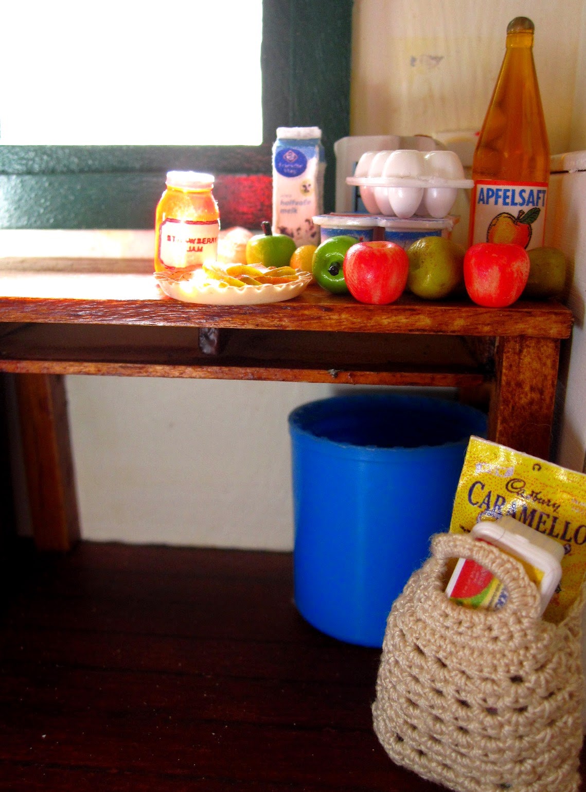 Inside of a miniature holiday house, showing a table with a selection of miniature grocery items, and a bag of groceries is on the floor underneath,