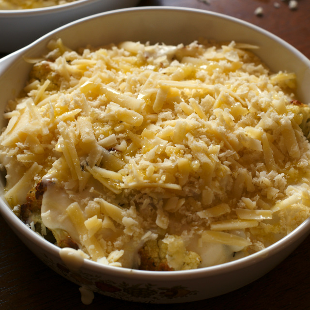 Roasted cauliflower topped with melted white cheese sauce  and shredded cheese in a vintage white casserole dish on a wooden background.