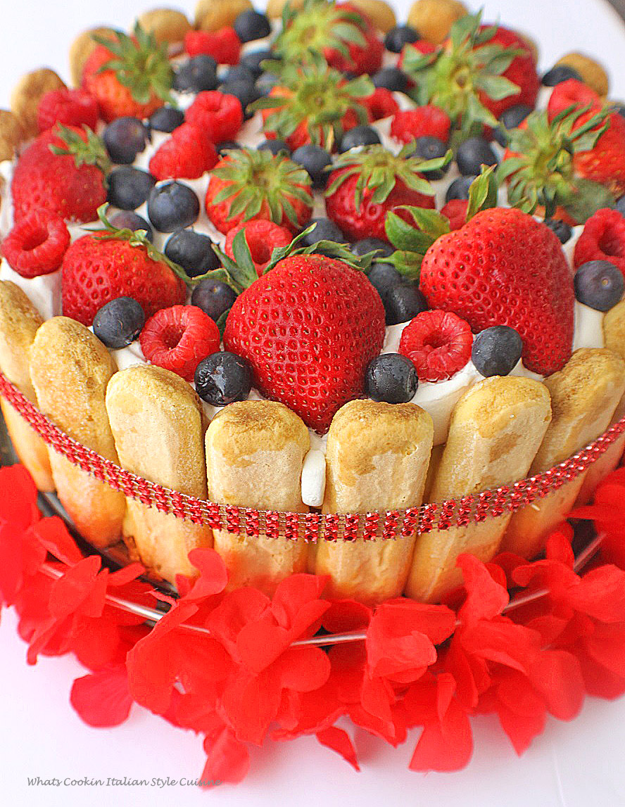 this is a layered cake trifle made in a springform pan. It has cake, lemon pudding, whipped cream, lady fingers around the edges and fresh raspberries, strawberries and blueberries on top. The cake is rich and elegant looking with a faux red diamond band all around and a Hawaiian lei around it on a red dish to serve it on