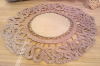 Table mat with a cloth centre and needleworked border. The border is made of serpentine shapes, and attached to the centre by fine beaded threads.