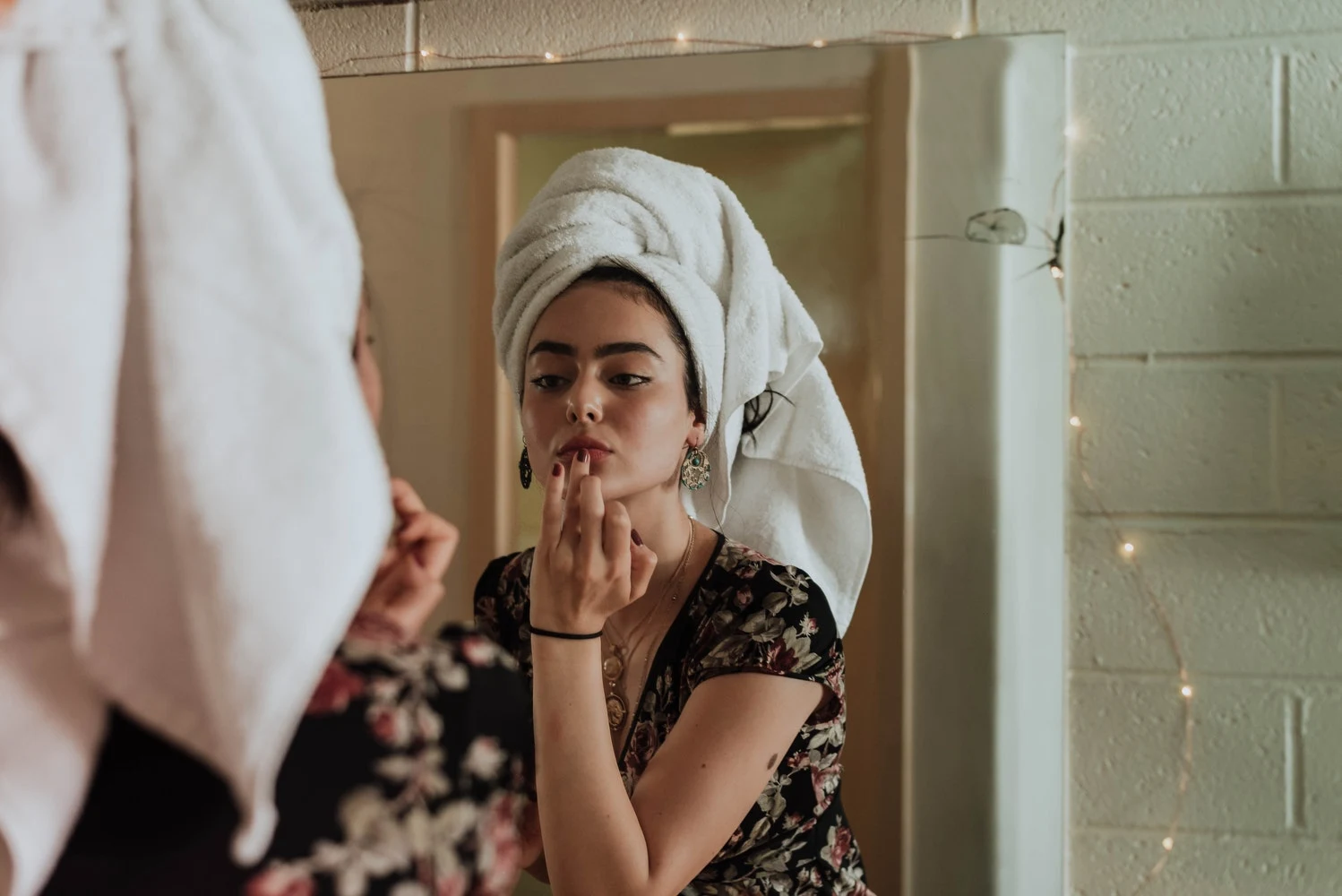 woman with a towel on her hair applies natural oil on to her face next to a mirror