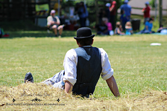 Player (a Man) sitting on the sidelines of the Vintage Baseball game waiting for his turn to bat. He is sitting on the grass and there are spectators across the field sitting on blankets and chairs.  The article is Vintage Baseball by Rosevine Cottage Girls