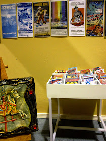 Modern miniature comic book store, interior view showing a rack of comics, a wall of posters and a fantasy art piece on an easel.