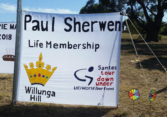 The banner looks home made on white fabric with black text painted on it.  A yellow crown depicting the "King Stage" is on the bottom left hand corner. A painted version of the Santos Tour Down Under logo is on the bottom right.  The banner is staked to the corner of a block of land of lawn and trees which is a reserve.