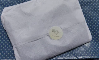 The bib wrapped up in white tissue paper sealed with a sticker with the Messy Me logo on, on top of the folded up messy mat.