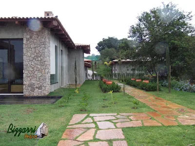 Execução do caminho de pedra no jardim com pedra Goiás tipo cacão assentada com juntas de grama, execução do revestimento de pedra rústica na fachada da residência em Piracaia-SP com os canteiros de viburno e os vasos de barro com as mudas de mexerica e o gramado com grama esmeralda.