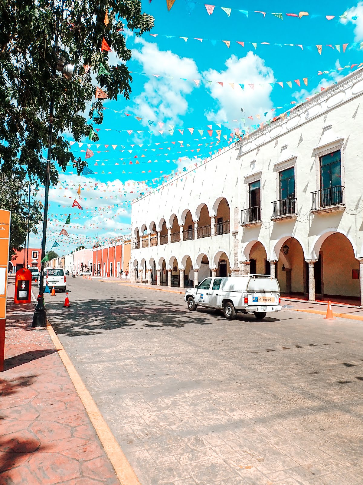 rue de la ville de valladolid au mexique, une voiture blanche roule sur la route d'autres à gauche sont garées on aperçoit les façades colorées et l'architecture typique de valladolid et des guirlandes de papiers colorés accrochés en l'air