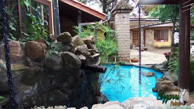 Detalhe da varanda do restaurante Recanto das Pedras com o lago de carpas com pedras ornamentais com o fundo do lago pintado de azul e nas fachadas as paredes de pedra com os pilares de pedra com o pergolado de madeira.