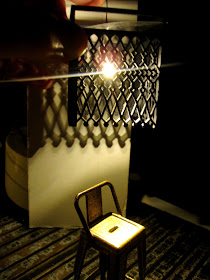 Modern dolls' house miniature scene in progress, with a copper-coloured cafe bar stool in the dark with a geometric-design light shade above, casting patterns on the wall.