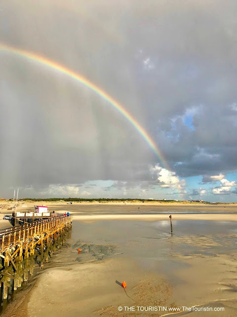 Two people walking on a wooden pier over the sea at low tide, next to a white sandy beach under a dark sky with a rainbow.