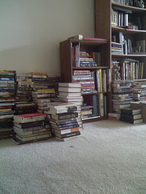 piles of books on the floor, next to full bookshelves