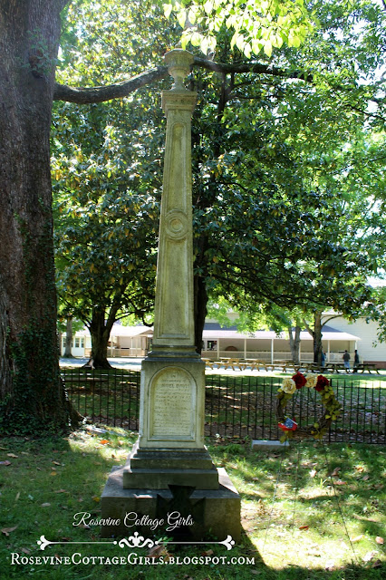 Photo of the very tall memorial stone for the grave of Sam Davis at the Sam Davis Home Cemetery in Tennessee.  The stone that was once white has green on it from sitting under the shade of the trees. It has a memorial statement on it but you can't read it in this photo as you are too far away.  The cemetery is surrounded by a rod iron fence and there is a pretty floral wreath next to the memorial. by RosevineCottageGirls.com