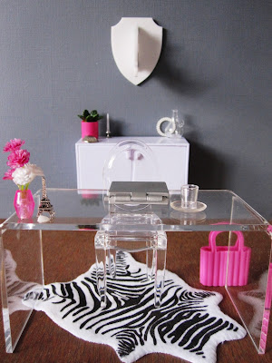 One-twelfth scale modern miniature office in shades of grey, white and hot pink. On the floor is a zebra-skin rug, and on the wall is a perspex rhino trophy head.
