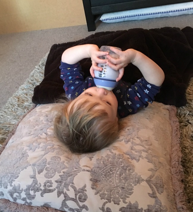 toddler lying on floor with head on cushion, blanket over lap, drinking milk from bottle