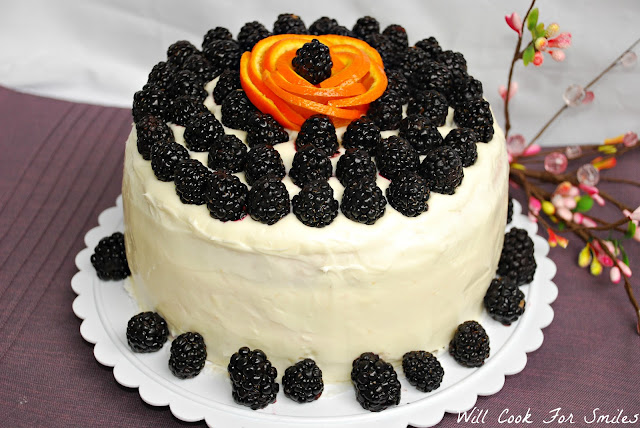 whole cake on a cake platter with white frosting and blackberries all around the bottom and blackberries all over the top with a orange peel made to look like a flower with a blackberry in the middle 