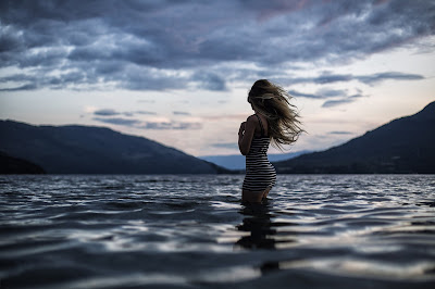 Chica rubia vestida metida en un lago con montañas de fondo