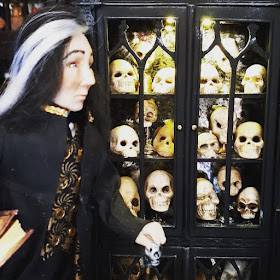 One-twelfth scale doll with black and grey hair in front of a cabinet full of skulls